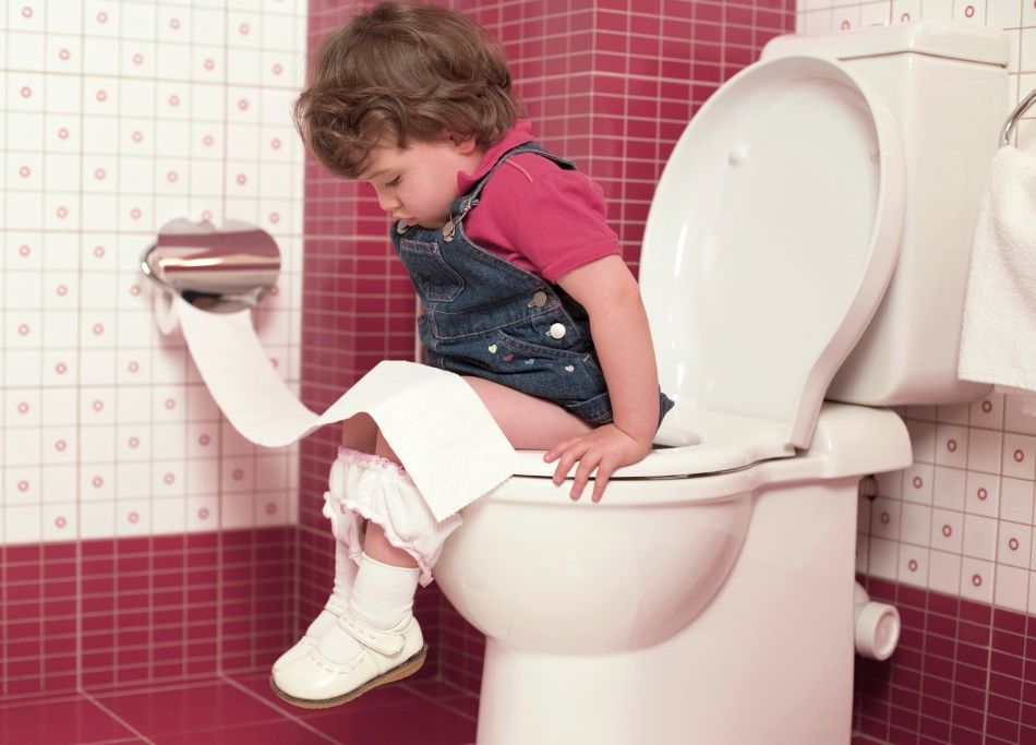 Ein Kind, das auf einer Toilette sitzt und Toilettenpapier über die Beine gerollt hat, während es sich mit den Händen am Sitz abstützt.