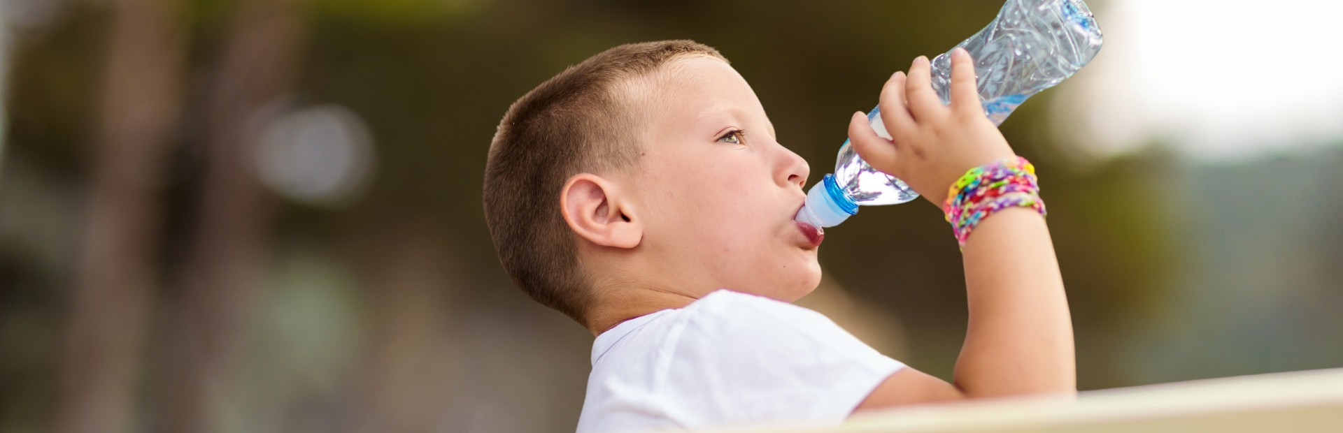 Ein Junge sitzt auf einer Bank und wird von der Seite beim Trinken aus einer Plastik-Wasserflasche fotografiert.
