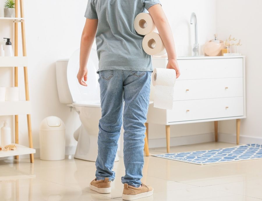 Ein Junge, den man von hinten sieht, steht im Badezimmer vor der Toilette mit einigen Rollen Toilettenpapier in der Hand und unter dem Arm.